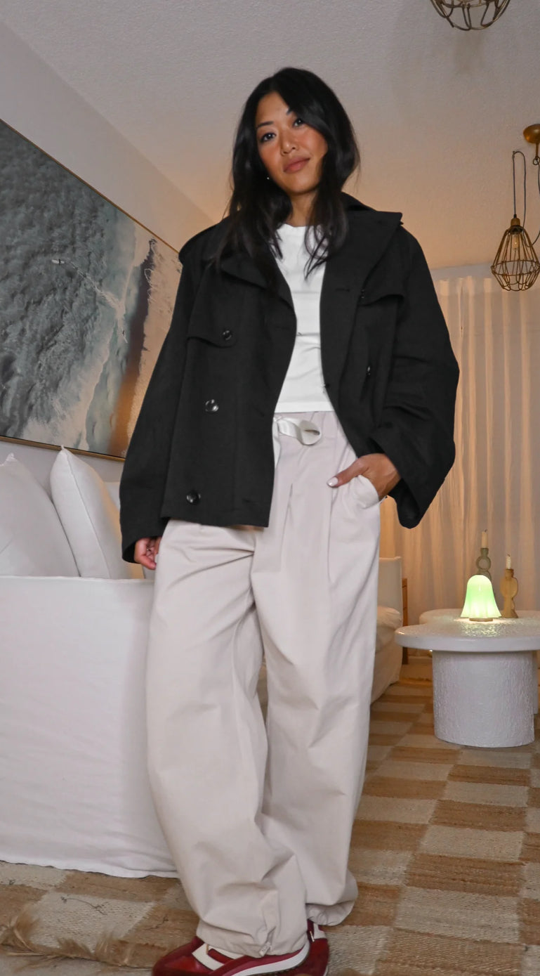 Woman in black jacket and white pants standing in a living room.