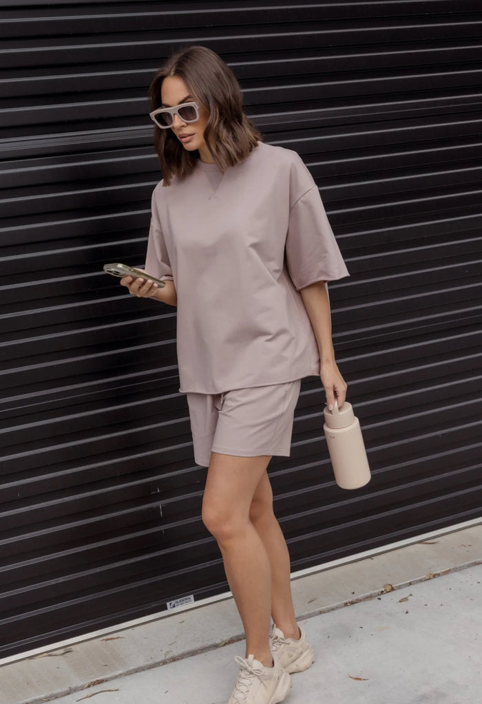 Woman in a beige outfit standing against a black wall.