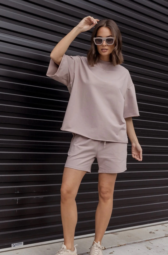 Woman wearing a light gray outfit with sunglasses against a black slatted wall.
