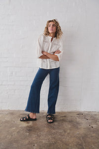 Person wearing a white patterned shirt and blue jeans standing against a white brick wall.