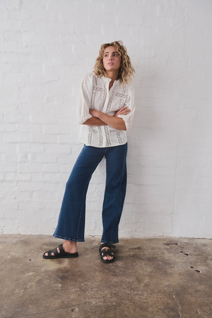 Person wearing a white patterned shirt and blue jeans standing against a white brick wall.