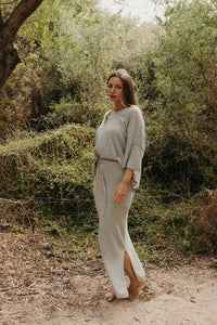 Woman in a gray outfit standing in a natural setting with greenery.